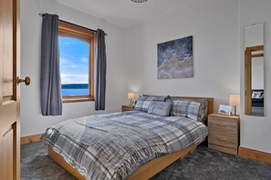 A bedroom with a bed, curtains, a nightstand, a lamp, a painting, and a view of the outdoors. - Cottage by Moray Firth With Stunning Views (Avoch)
