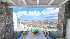 Un espace de restauration extérieur aux murs de pierre, avec une table dressée pour deux personnes, offrant une vue sur un paysage côtier, une piscine et la mer.