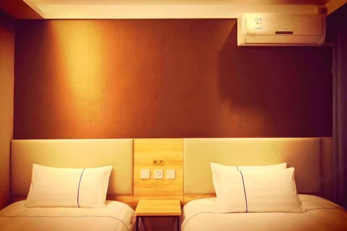 A hotel room with two beds, a wooden headboard, a small bedside table, and an air conditioning unit.