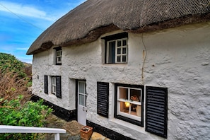 A traditional thatched-roof house with white walls and black shutters, featuring multiple windows and a visible door. - Cottage in Sennen Cove With Stunning Sea Views (Penzance)