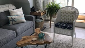Ett modernt vardagsrum med en grå soffa, ett soffbord i trä, en fåtölj i rotting och ett stort fönster med gardiner.