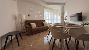 A living room with a brown sofa, a dining table with chairs, a TV mounted on the wall, and a lamp. - Láttic, Terres de lébre Biosphere Reserve (La Ràpita)