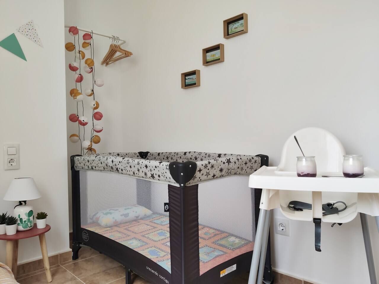 Een kinderbed met stapelbed en een sterrenpatroon bovenin, een klein tafeltje met een lamp, en een witte stoel met twee potjes erop.