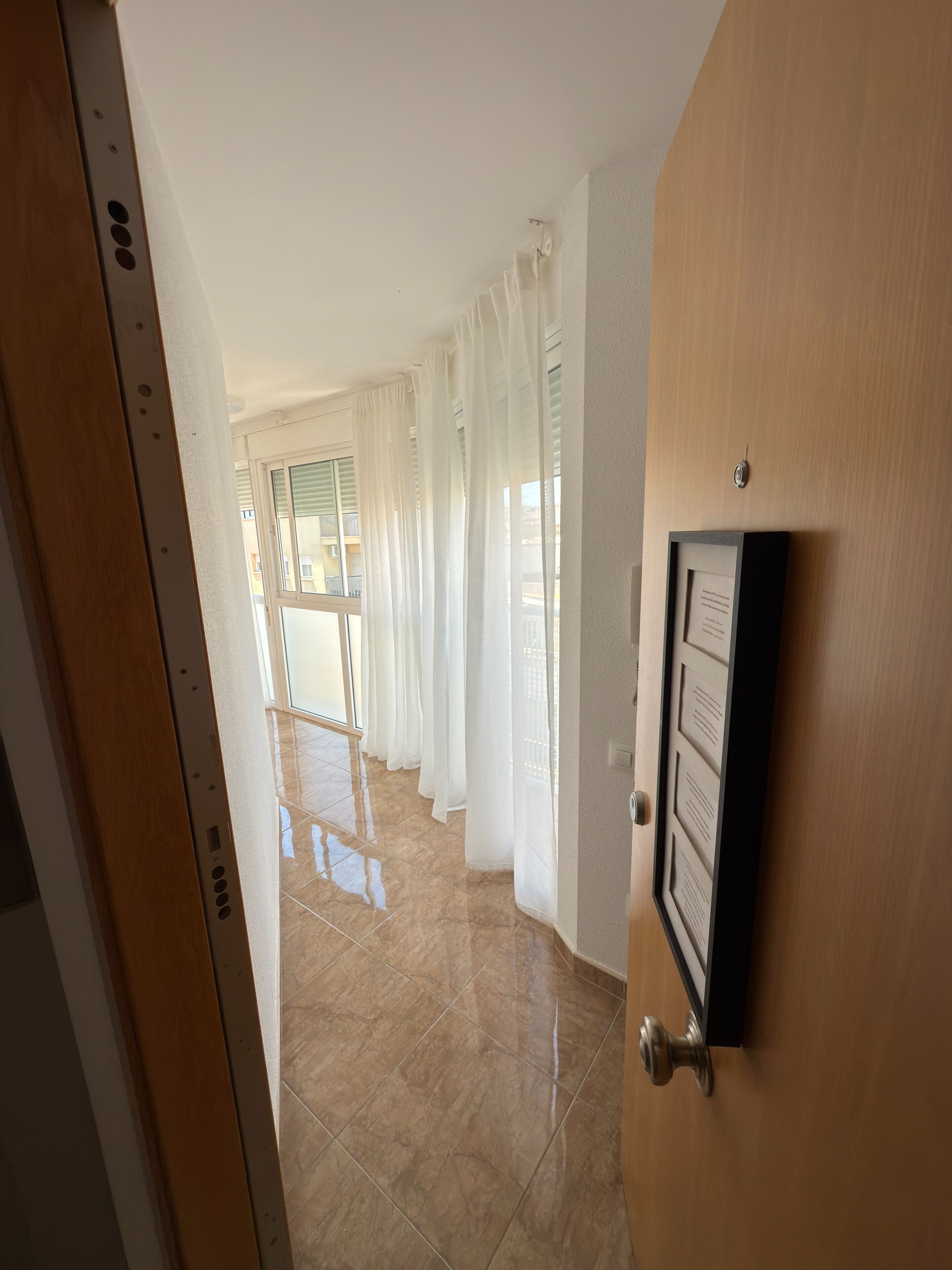 Un pasillo con puerta de madera, suelo de baldosas y cortinas blancas.