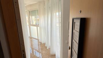 Un pasillo con puerta de madera, suelo de baldosas y cortinas blancas.