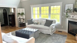 A living room with a white sofa, grey cushions, a grey rug, and a fireplace. There is a window with a view of trees, and a bookshelf with books and decorative items. - Beautiful, private getaway, close to all New England attractions (Hampton Falls)