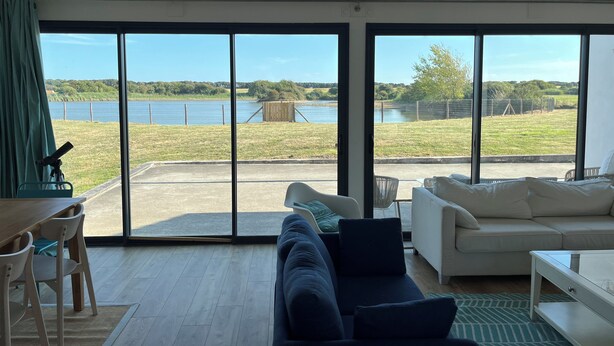 Un salon moderne doté d’une grande porte-fenêtre coulissante donnant sur un balcon avec vue sur un lac et la verdure environnante.