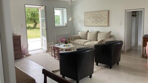 Un salon spacieux avec un canapé blanc, des fauteuils noirs et une table à manger. Une grande fenêtre laisse entrer la lumière naturelle et offre une vue sur la verdure environnante.