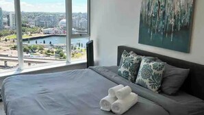 Ein Schlafzimmer mit einem großen Bett, einem Gemälde an der Wand und Blick auf eine Stadtkulisse durch ein großes Fenster.