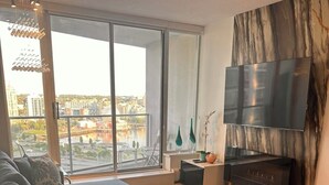 Ein modernes Wohnzimmer mit einer großen Couch, einem kleinen Tisch, einem Flachbildfernseher und Blick auf die Stadt durch große Fenster.