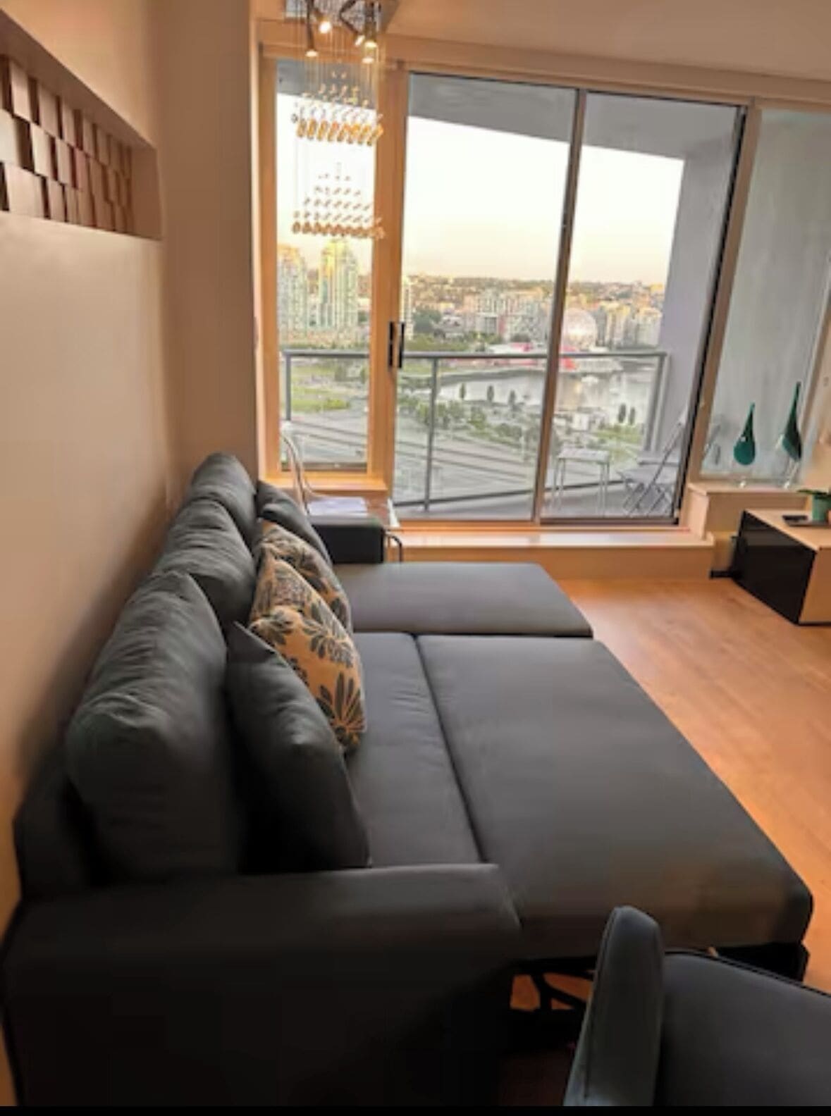 Una sala de estar moderna con un sofá gris, vistas de un paisaje urbano a través de amplios ventanales y una lámpara decorativa.