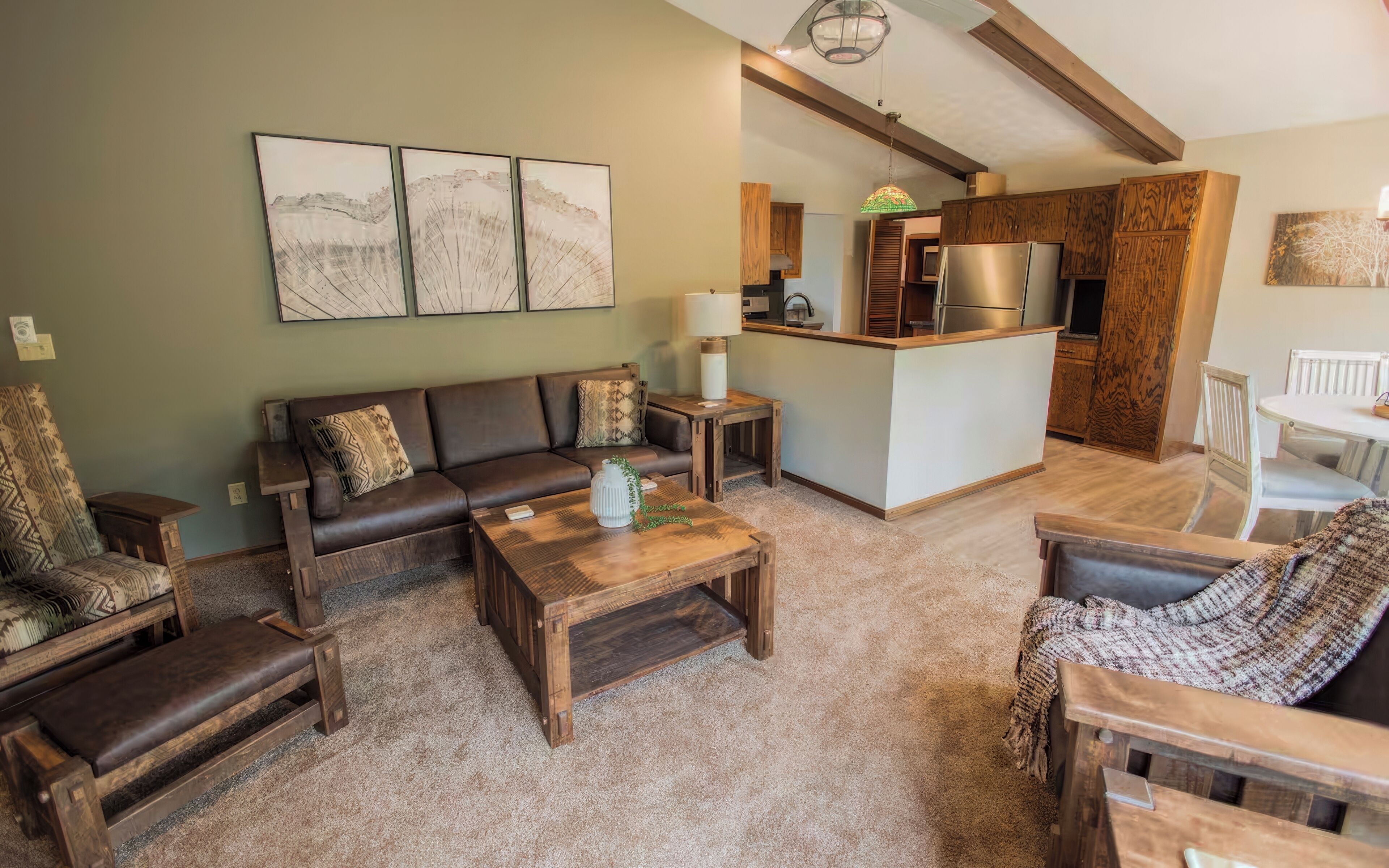 A living room with a brown leather sofa, wooden coffee table, and patterned armchairs. A kitchen area with wooden cabinets and a refrigerator is visible in the background.