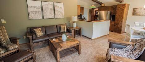 A living room with a brown leather sofa, wooden coffee table, and patterned armchairs. A kitchen area with wooden cabinets and a refrigerator is visible in the background.