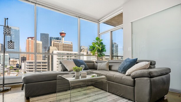 A modern living room with a sectional sofa, a glass coffee table, and a view of a cityscape through large windows. - Downtown Chic Retreat | Elegant + Free Parking (Calgary)