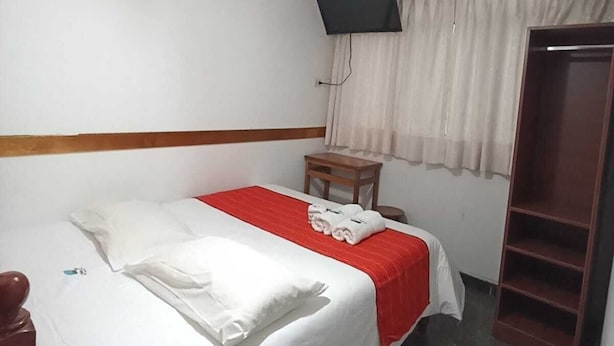 A hotel room with a bed, a television mounted on the wall, a small table, and a wardrobe.