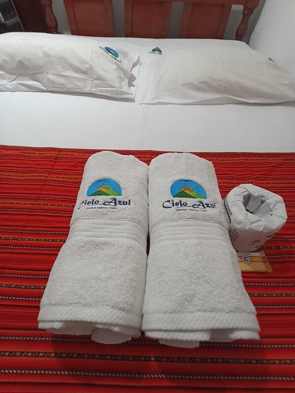 Cama com almofadas brancas e duas toalhas enroladas com a marca 'Cielo Azul' em um edredom listrado de vermelho.
