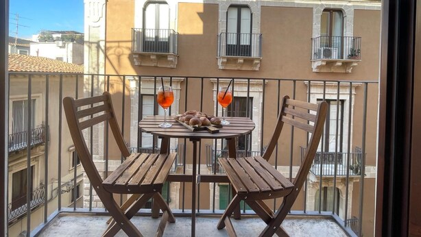 A balcony with two wooden chairs and a table, two drinks, and a plate of pastries. - Maison Etnea (Catania)