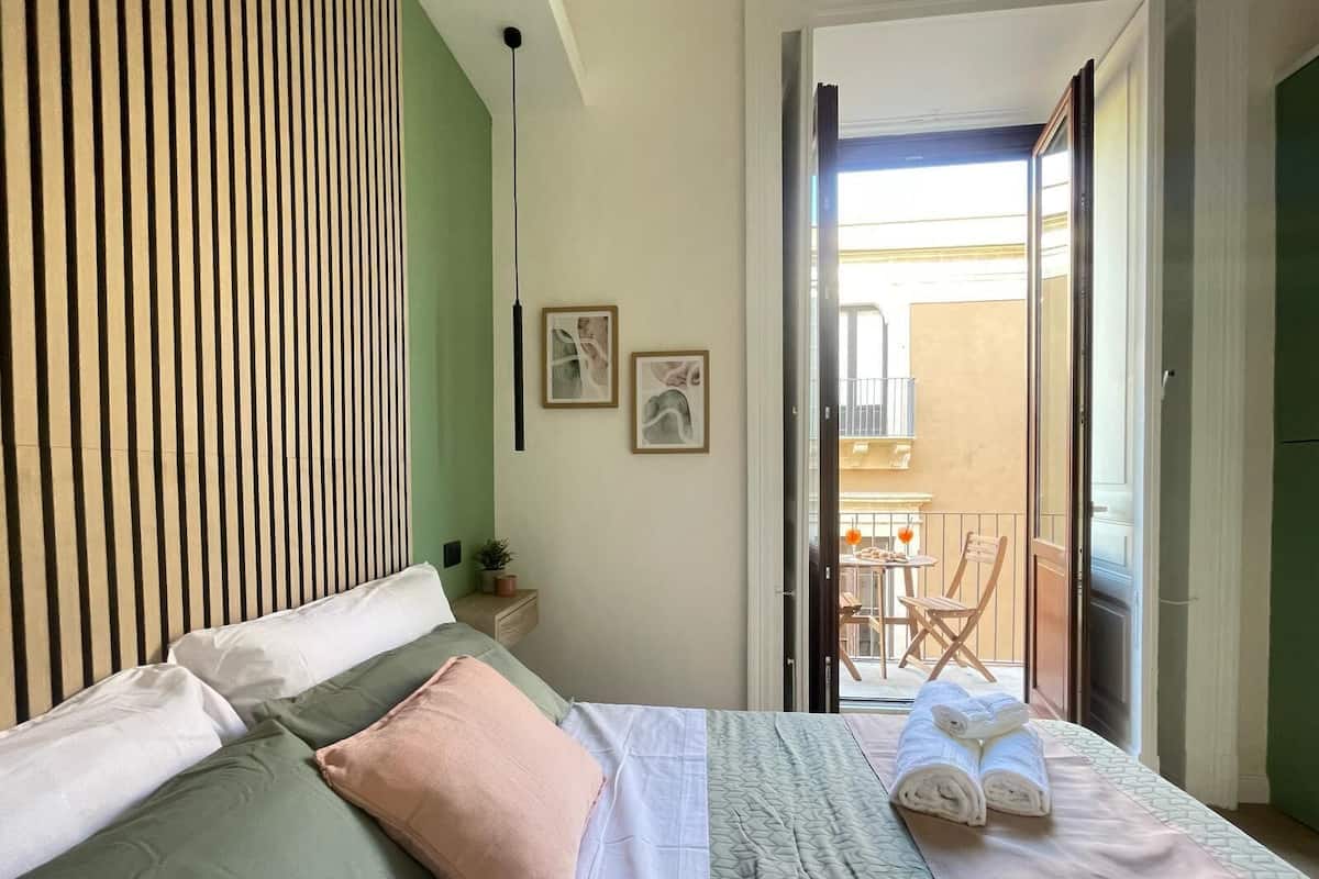 A bedroom with a bed, pillows, a bedside table with a plant, and a view of an outdoor balcony with a table and chairs.