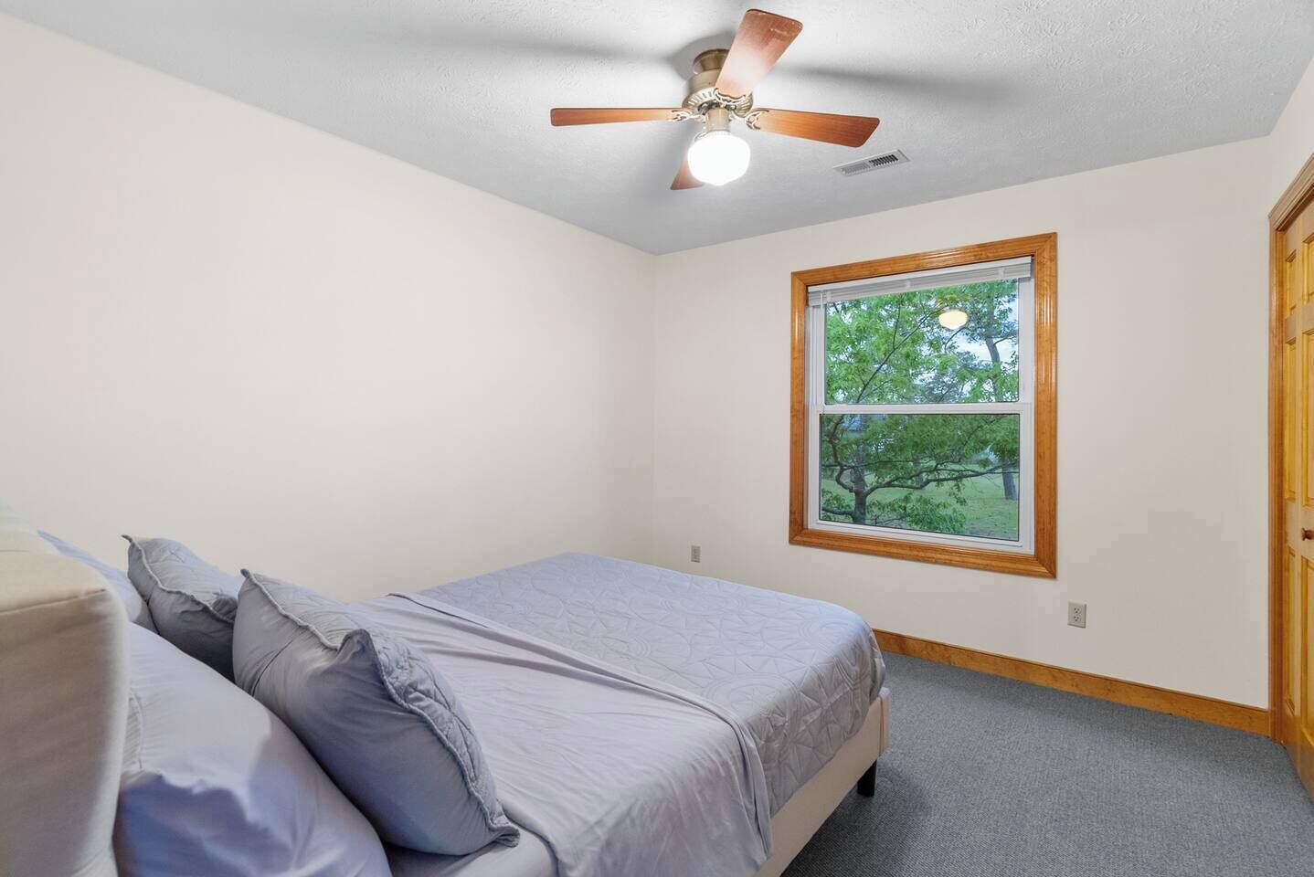 Une chambre à coucher avec un lit, des oreillers, un ventilateur de plafond, une fenêtre donnant sur des arbres et une porte.