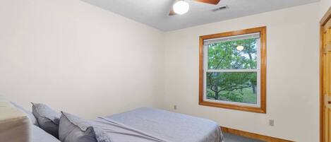 Une chambre à coucher avec un lit, des oreillers, un ventilateur de plafond, une fenêtre donnant sur des arbres et une porte.