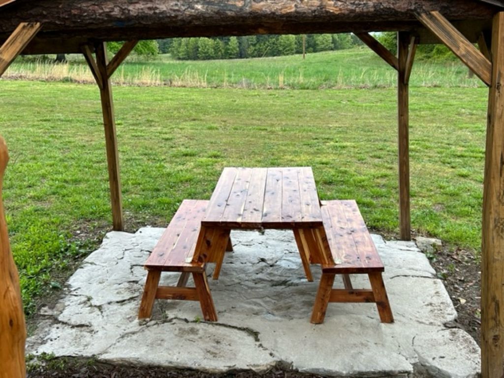 Una mesa de picnic de madera con bancos bajo un techo de madera en un campo cubierto de hierba.