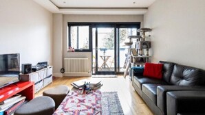 A modern living room with a black leather sofa, a red entertainment unit, and a large window with a view of the outdoors. - Elegant London Escape — 2BR Apartment with Breakfast & Daily Cleaning (London)