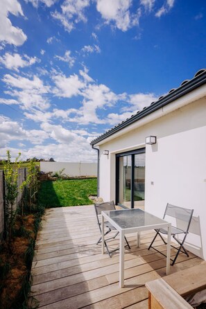 A modern outdoor patio with a glass table and chairs, a wooden deck, and a white building with a black roof.