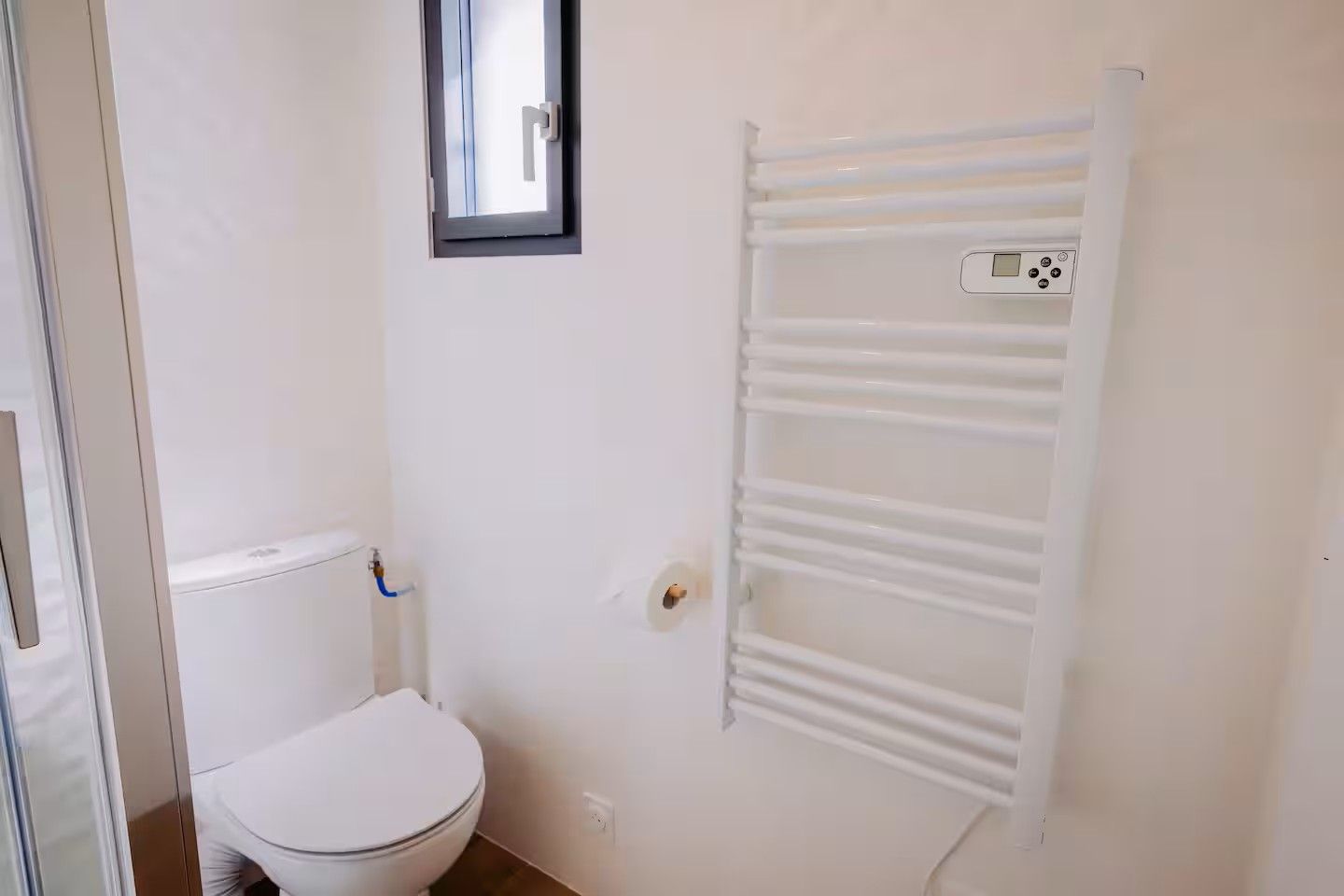 Une petite salle de bain blanche, avec des toilettes, un porte-serviettes blanc et une fenêtre située au-dessus des toilettes.