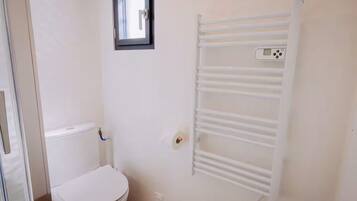 Une petite salle de bain blanche, avec des toilettes, un porte-serviettes blanc et une fenêtre située au-dessus des toilettes.