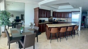 A modern kitchen with dark wood cabinets, a granite countertop, and a large island with bar stools. - Las Olas Grand 3br Luxury- Beach, Pools, Hot tubs (Rosarito)