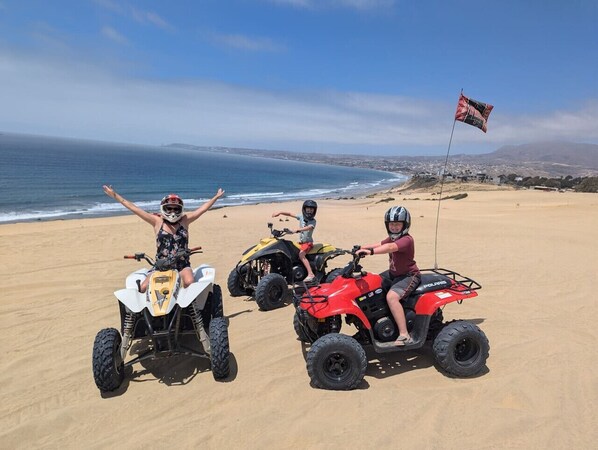 Tres personas en vehículos todo terreno sobre una playa arenosa, con una bandera al fondo.