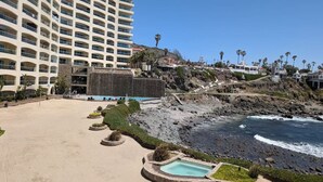A beachfront hotel with a pool area, a rocky shoreline, and a large building in the background. - Las Olas Grand 3br Luxury- Beach, Pools, Hot tubs (Rosarito)