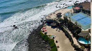 An aerial view of a coastal hotel with a rocky shoreline, a beach area with sun loungers, a tennis court, and a pool. - Las Olas Grand 3br Luxury- Beach, Pools, Hot tubs (Rosarito)