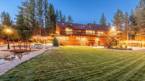 Un grand chalet à étages, doté d’un éclairage extérieur soigné, d’un espace de détente extérieur abrité et d’un petit ruisseau en premier plan.