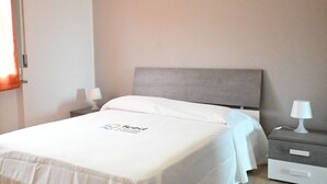 Une chambre d’hôtel avec un lit, deux tables de chevet avec des lampes, une tête de lit grise et un logo sur la couvre-lit.