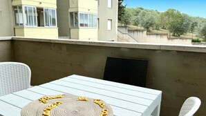 Un balcon avec une table et des chaises blanches, un set de table en fibres tressées et une vue sur un immeuble et des arbres.