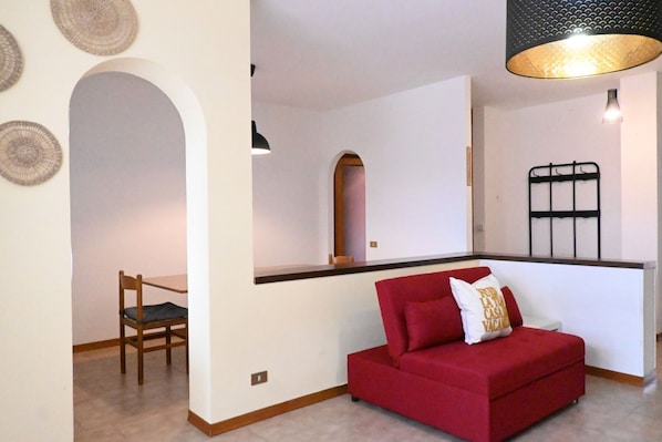 Un canapé rouge avec un coussin, une chaise en bois, une étagère fixée au mur et un luminaire suspendu.