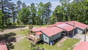 Une maison à deux étages avec un toit rouge, une terrasse en bois et une vue sur un lac entouré d’arbres.
