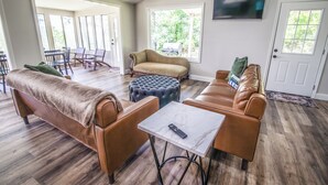 Un salon spacieux avec un canapé en cuir marron, un pouf assorti et une table basse blanche dotée d’une télécommande. Un téléviseur est fixé au mur, et une grande fenêtre laisse entrer la lumière naturelle.