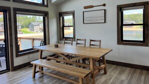 Un comedor con mesa y bancos de madera, ventanas amplias con vistas a un lago y edificios, y una cita enmarcada en la pared.