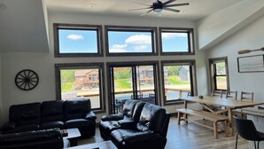 Amplia sala de estar con un sofá de cuero negro, una mesa de centro de madera y un comedor con mesa y sillas de madera. Amplias ventanas ofrecen vistas al exterior.