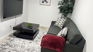 Ein modernes Wohnzimmer mit einem an der Wand befestigten Flachbildfernseher, einem Couchtisch mit einer Pflanze, einem grauen Sofa mit gemusterten Kissen und einer großen Topfpflanze.