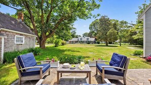 A patio with two blue cushioned chairs, a small table with a pitcher of lemonade, and a view of a large house in the background.