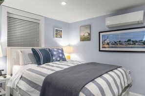 A bedroom with a bed, a nightstand, a lamp, and a window with blinds. - Pet-Friendly Beachside Condo + Parking on Comm St (Provincetown)