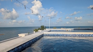 A pool area with clear blue water, a tiled deck, and a view of the sea under a partly cloudy sky. - Modern and brand new 3-bedroom condo with private rooftop pool  with majestic views. (Caye Caulker)