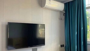 A room with a mounted flat-screen TV, a window with teal curtains, and an air conditioning unit. - Shuhao Hotel (Shanghai)