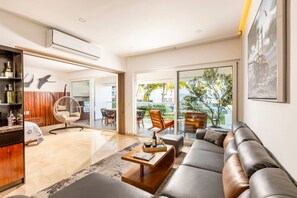 Uma sala de estar moderna com sofá, mesa de centro e cadeiras, uma estante de madeira e uma janela grande com vista para o verde.
