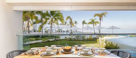 Mesa de comedor servida con platos, tazones y cubiertos, con vista a un área junto a la piscina con palmeras y sombrillas.