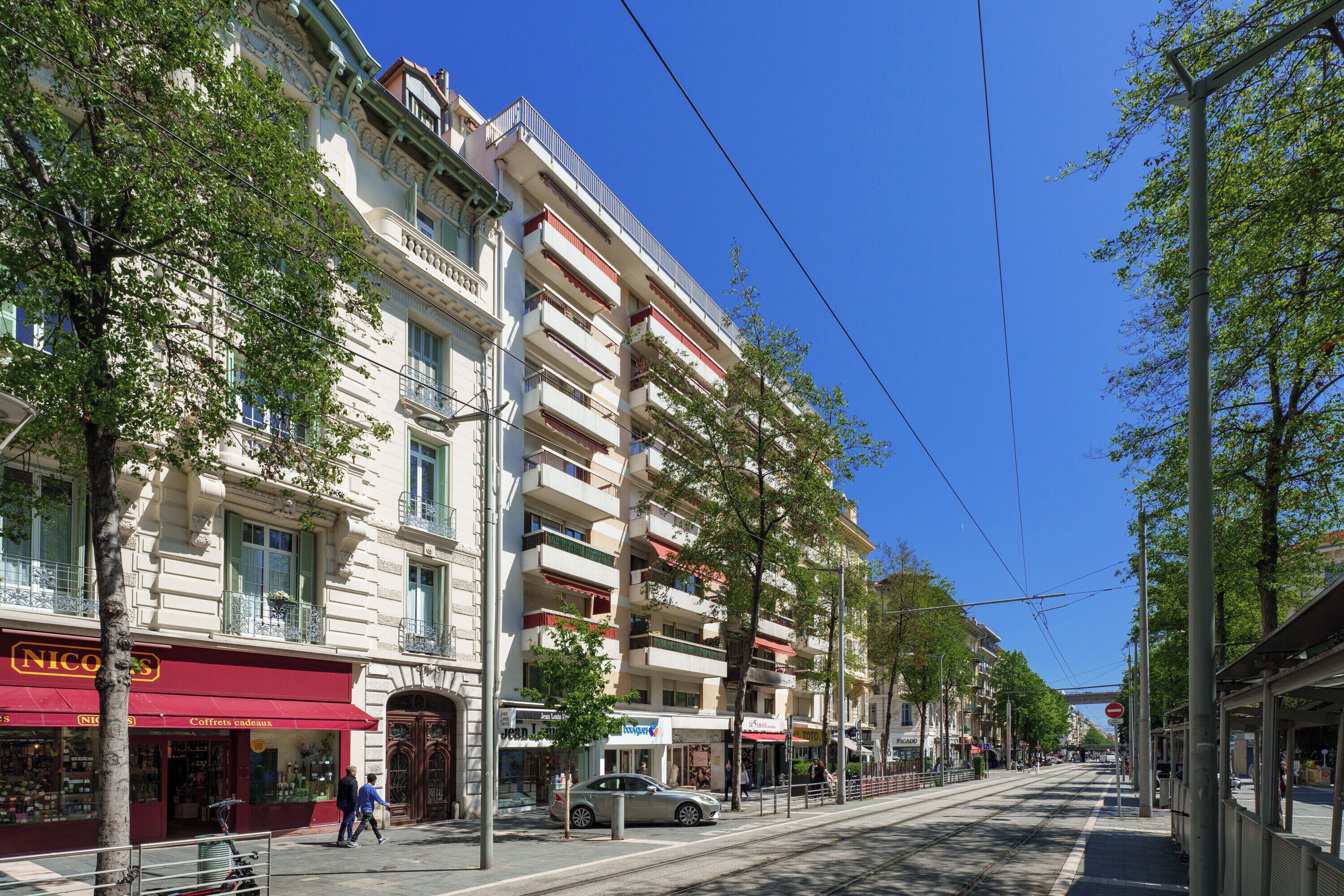 A multi-story building with balconies, a red awning with the word "NICOLES," and a tram line running down the street.