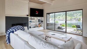 A modern living room with a white sofa, a fireplace, a Netflix TV, and a large sliding glass door leading to a balcony with a lake view. - Cozy 3BR by the lagoon w/ hot tub (Stinson Beach)